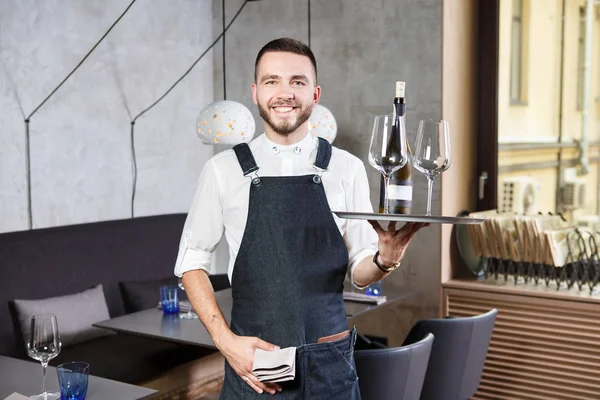 Ένα νεαρό, όμορφος Καυκάσιος σερβιτόρος στέκεται στο εσωτερικό του εστιατορίου με ένα δίσκο στο χέρι, δύο ποτήρια και μια ζαριά του κρασιού. Ντυμένος με στολή ποδιά και πουκάμισο. Το θέμα εξυπηρέτηση — Φωτογραφία Αρχείου