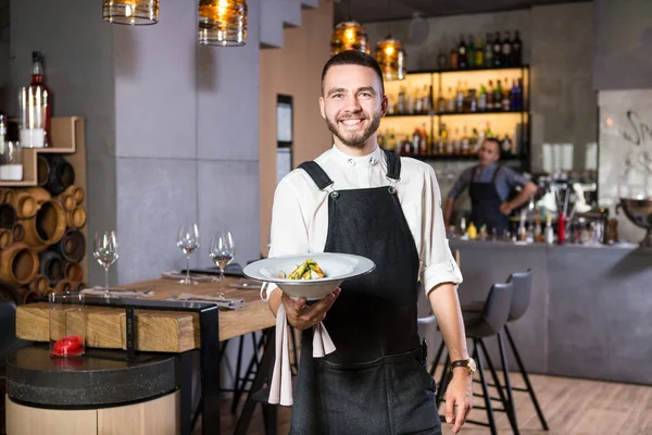 A handsome young guy with a beard dressed in an apron standing in a restaurant and holding a white plate with a moth. Against the background, the bar counter and the loft style interior — Stock Photo, Image