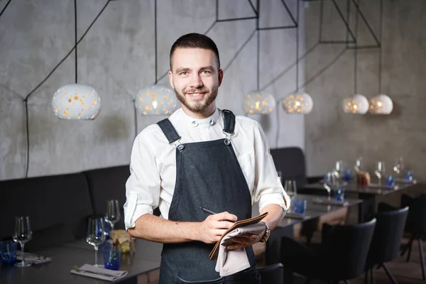 A young, smiling waiter in a restaurant, standing next to the tables with a glass of wine. Dressed in an apron, will take an order holding a notebook and pen — Stock Photo, Image