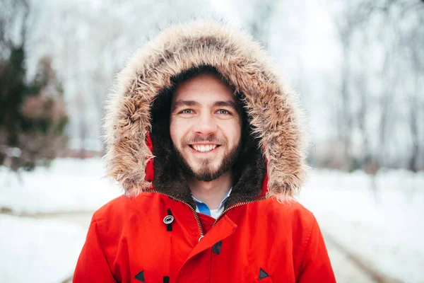 Portrait, close-up of a young stylishly dressed man smiling with a beard dressed in a red winter jacket with a hood and fur on his head. Winter and frost theme. — Stock Photo, Image