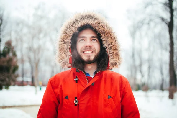 Retrato, close-up de um jovem elegantemente vestido homem sorrindo com uma barba vestida com um casaco de inverno vermelho com um capuz e pêlo na cabeça. Inverno e tema de geada . — Fotografia de Stock