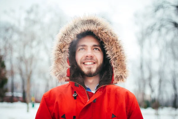 Retrato, close-up de um jovem elegantemente vestido homem sorrindo com uma barba vestida com um casaco de inverno vermelho com um capuz e pêlo na cabeça. Inverno e tema de geada . — Fotografia de Stock