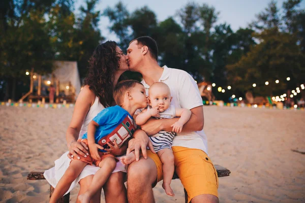 The concept of a family vacation. Young family sitting on a bench in the evening on a sandy beach. Mom and Dad kiss, the older brother kisses the younger on the cheek.