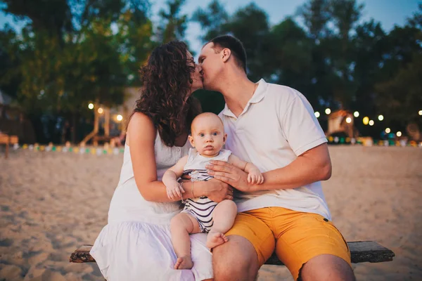 Tema aile tatil yaz Nehri yakınında. erkek ve kadın, elindeki sürpriz komik bir duygu ile oturan bir küçük çocuk evlat öp. Gün batımında sahilde akşamları — Stok fotoğraf