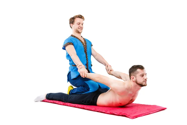 Un joven hombre guapo médico masajista en un traje azul hace una pose tradicional de masaje takia a un tipo acostado en una vida en una alfombra de alfombra. Fondo blanco aislado. Tema Masaje tailandés — Foto de Stock