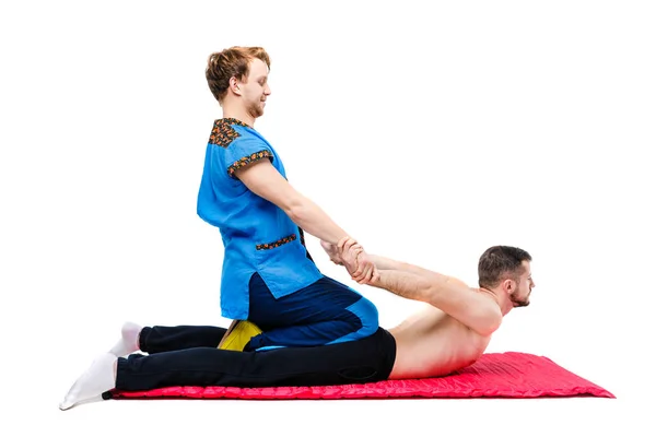 Un joven hombre guapo médico masajista en un traje azul hace una pose tradicional de masaje takia a un tipo acostado en una vida en una alfombra de alfombra. Fondo blanco aislado. Tema Masaje tailandés — Foto de Stock