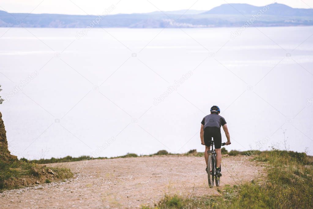 Theme tourism and cycling on mountain biking. young guy riding down at high speed on rocky, mountain road backdrop Mediterranean Sea in Spain on shore of kosta brava in helmet and bicycle shorts