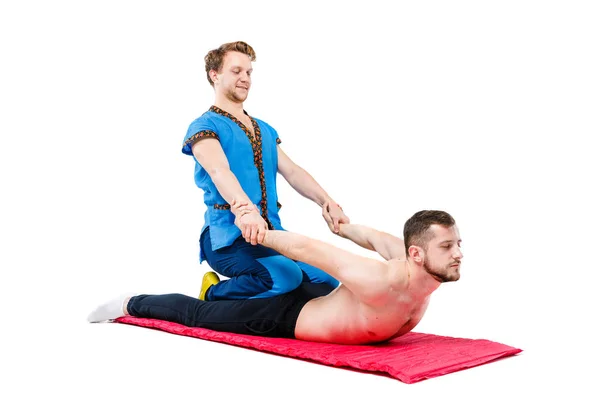 Un joven hombre guapo médico masajista en un traje azul hace una pose tradicional de masaje takia a un tipo acostado en una vida en una alfombra de alfombra. Fondo blanco aislado. Tema Masaje tailandés — Foto de Stock