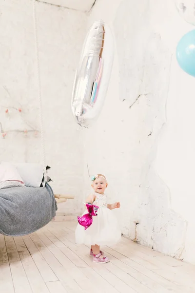 Sujeito crianças aniversário é crianças de um ano. Menina pequena na sala brilhante contra a parede branca no vestido e na bola inflável segurando loira na forma de unidade, ao lado do grupo de bolas cores diferentes — Fotografia de Stock