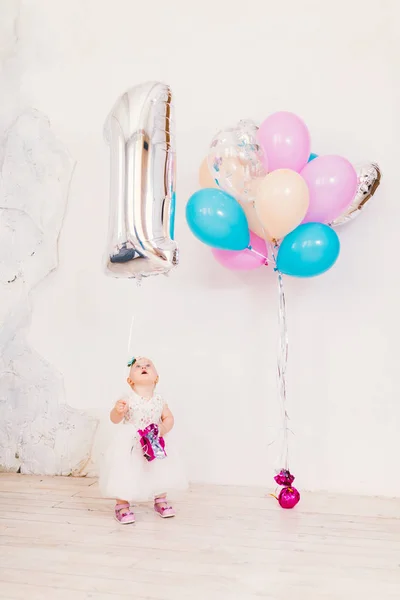 Субъект детского дня рождения один год. Маленькая девочка в яркой комнате против белой стены в платье и блондинка проведение надувной мяч в виде единицы, рядом с группой шаров различных цветов — стоковое фото