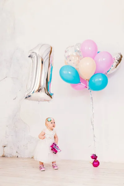 Субъект детского дня рождения один год. Маленькая девочка в яркой комнате против белой стены в платье и блондинка проведение надувной мяч в виде единицы, рядом с группой шаров различных цветов — стоковое фото