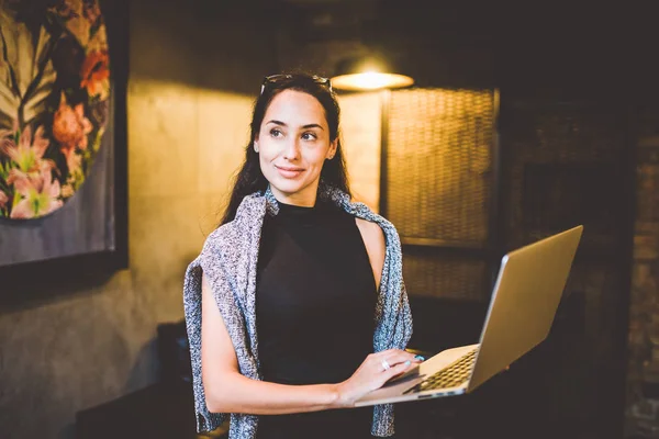 O conceito de pequenas empresas e tecnologia. Jovem mulher de negócios bonita morena em vestido preto e camisola cinza fica na cafeteria com laptop nas mãos. Nos óculos de cabeça para a vista — Fotografia de Stock