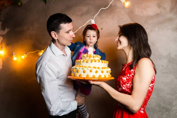 Tema familjesemester barnens födelsedag och blåser ut ljusen på stor tårta. ung familj tre människor stående och anläggning 5-åriga dotter i gården mot grå vägg och krans gul lökar — Stockfoto