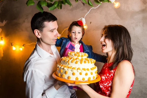Тема сімейного свята дитячий день народження і вибуху свічок на великому торті. молода сім'я з трьох чоловік стоїть і тримає п'ятирічну дочку на подвір'ї проти сірої стіни і жовтих цибулин гірлянди — стокове фото