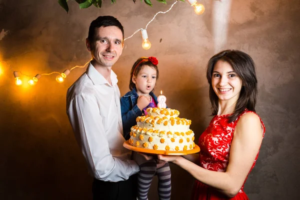 Θέμα οικογενειακές διακοπές Παιδικά γενέθλια και σβήσιμο κεριών σε μεγάλη τούρτα. νεαρή οικογένεια από τρεις άνθρωποι στέκεται και εκμετάλλευση 5 χρονών κόρη στο ναυπηγείο γκρίζο τοίχο και γιρλάντα κίτρινους βολβούς — Φωτογραφία Αρχείου