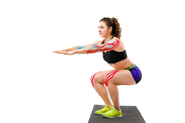 Reabilitação de fita cinesiologia tema e saúde de atletas. Menina bonita fazendo um exercício de agachamento em um tapete preto em um fundo isolado branco. No braço e joelho, uma fita colorida pegajosa — Fotografia de Stock