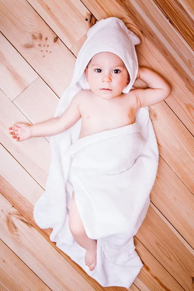 Αγοράκι με καστανά μάτια είναι πέντε μηνών τυλιγμένο σε μια λευκή πετσέτα με τα αυτιά σε φόντο ξύλινη . — Φωτογραφία Αρχείου
