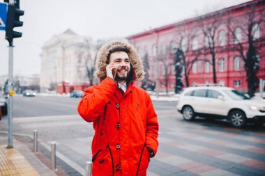 Sakal ve kırmızı ceket başlıklı bir öğrenci genç adam cep telefonu kullanır, üniversite veya kolej bina kırmızı zemin üzerine gülümseme ile telefonda konuşurken yakınındaki baş, elinde tutan