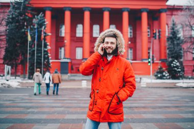 Sakal ve kırmızı ceket başlıklı bir öğrenci genç adam cep telefonu kullanır, üniversite veya kolej bina kırmızı zemin üzerine gülümseme ile telefonda konuşurken yakınındaki baş, elinde tutan