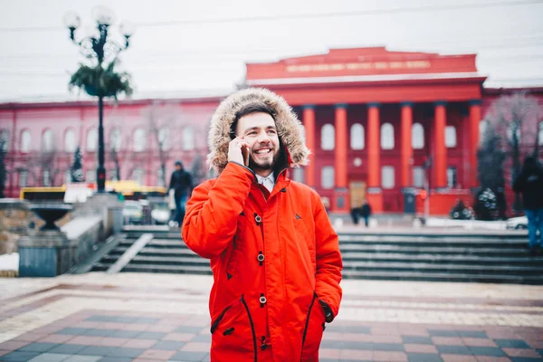 ひげと赤いジャケット フード学生で若い男が携帯電話を使用して、頭の近くの彼の手を保持する赤い大学やカレッジの建物の背景に笑みを浮かべて、電話で話しています。 — ストック写真