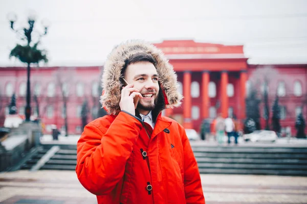 젊은 남자 수염과 빨간 자 켓 후드 학생 휴대 전화를 사용 하 여, 그의 손을 머리 근처에 레드 대학 또는 대학 건물의 배경에 미소와 전화 통화 — 스톡 사진
