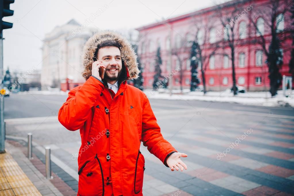 young guy with beard and red jacket in hood a student uses mobile phone, holds in his hand near the head, talking on the phone with smile on the background of red building the university or college