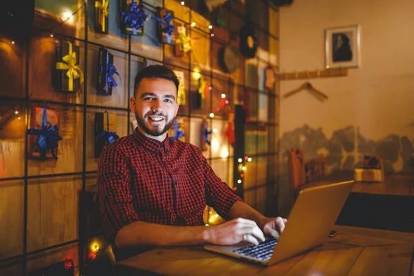 テーマ トレーニングとコンピューター。ひげとシャツの若い男は、夕方には木製のテーブルでコーヒー ショップでキーボードのプリント、ノート パソコンを使用します。クリスマスの装飾とハング花輪 — ストック写真