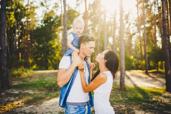 Família mãe, pai e filha se senta no papai nos ombros, e os pais se beijam na natureza na floresta no verão ao pôr do sol — Fotografia de Stock