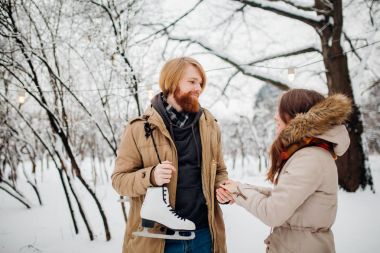 Winter and date. Young couple, lovers man and woman in winter on a background of snow and forest holding hands and smiling. A guy with long hair and a beard is holding skates and looking at the girl clipart