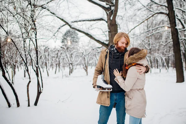 Зима и дата. Молодая пара влюблена в мужчину и женщину зимой на фоне заснеженных деревьев в парке обнимаются. Парень с длинными волосами и бородой держит коньки и любит девушку. — стоковое фото