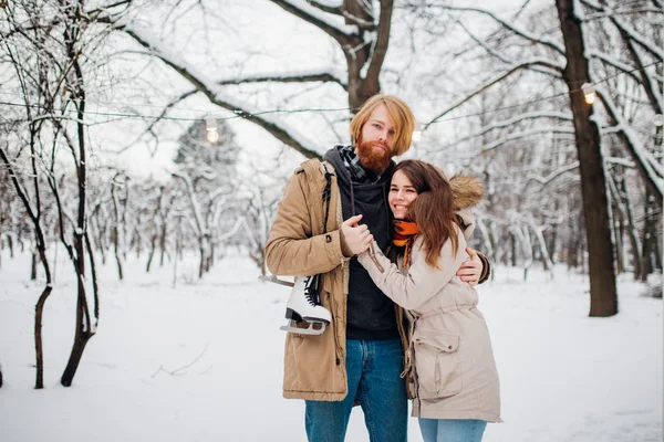 Hiver et rendez-vous. Jeune couple amoureux de l'homme et de la femme en hiver sur fond d'arbres enneigés dans le parc sont embrasser. Un gars avec les cheveux longs et la barbe garde les patins et aime petite amie — Photo