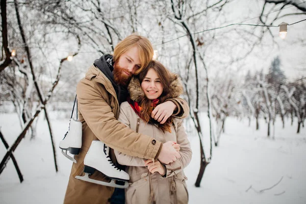 Зима и дата. Молодая пара влюблена в мужчину и женщину зимой на фоне заснеженных деревьев в парке обнимаются. Парень с длинными волосами и бородой держит коньки и любит девушку. — стоковое фото