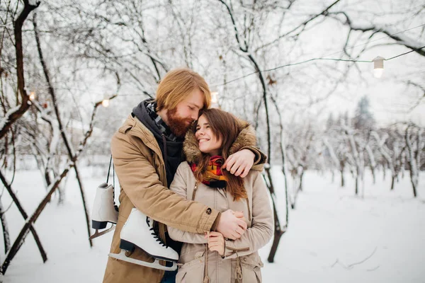 Χειμώνα και την ημερομηνία. Νεαρό ζευγάρι στην αγάπη με τον άνθρωπο και η γυναίκα το χειμώνα φόντο χιονισμένες δένδρα στο πάρκο είναι αγκαλιά. Ένας άντρας με μακριά μαλλιά και γενειάδα κρατά πατίνια και αγαπά τη φίλη — Φωτογραφία Αρχείου