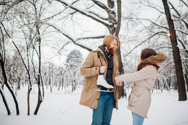 Зима и дата. Молодая пара, влюблённые мужчина и женщина зимой на фоне снега и леса держатся за руки и улыбаются. Парень с длинными волосами и бородой держит коньки и смотрит на девушку — стоковое фото