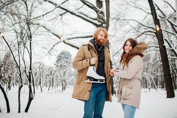 Hiver et rendez-vous. Jeune couple, amoureux homme et femme en hiver sur fond de neige et de forêt tenant la main et souriant. Un gars aux cheveux longs et à la barbe tient des patins et regarde la fille — Photo