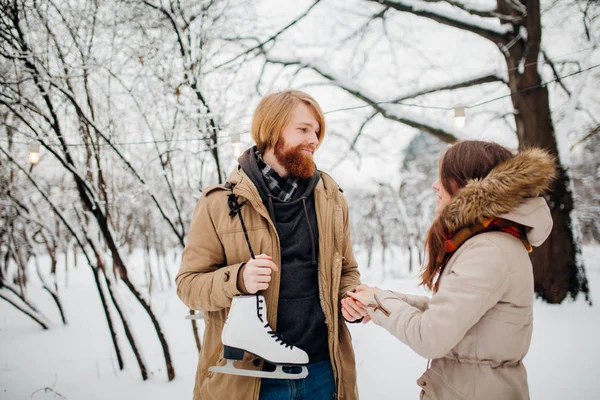 Hiver et rendez-vous. Jeune couple, amoureux homme et femme en hiver sur fond de neige et de forêt tenant la main et souriant. Un gars aux cheveux longs et à la barbe tient des patins et regarde la fille — Photo