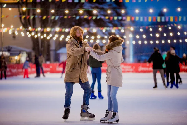 Thema Eisbahn und Liebespaar. Auf der von Glühbirnen und Lichtern beleuchteten Eisbahn in der Stadt treffen sich junge, stylische Menschen, die Hand in Hand in Menschenmenge fahren. Eislaufen im Winter zu Weihnachten in der Eisarena — Stockfoto