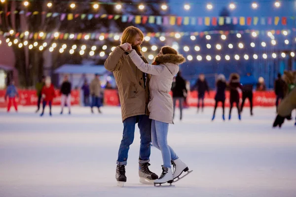 Thema Eisbahn und Liebespaar. Auf der von Glühbirnen und Lichtern beleuchteten Eisbahn in der Stadt treffen sich junge, stylische Menschen, die Hand in Hand in Menschenmenge fahren. Eislaufen im Winter zu Weihnachten in der Eisarena — Stockfoto