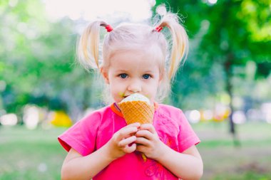 küçük kız sarışın bir gözleme Kupası parkta yeşil yaz zemin üzerine tatlı mavi dondurma yemek. onun yüz ve yanak ve gülüyor bulaşmış. Giymiş parlak şık giysiler