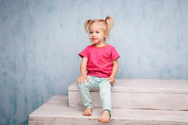 Grappige blauwogige meisje kind blonde met een kapsel twee extensie op haar hoofd zittend op een roddel op de achtergrond van een oude getextureerde muur in blauw. Gekleed in stijlvolle lichte kleding — Stockfoto
