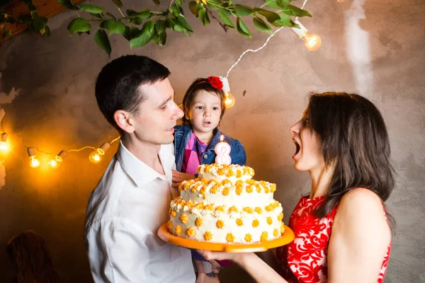 Theme family holiday childrens birthday and blowing out candles on large cake. young family of three people standing and holding 5 year old daughter in yard against gray wall and garland yellow bulbs