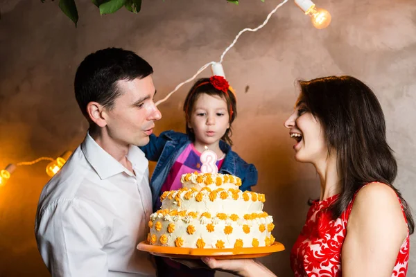 Theme family holiday childrens birthday and blowing out candles on large cake. young family of three people standing and holding 5 year old daughter in yard against gray wall and garland yellow bulbs