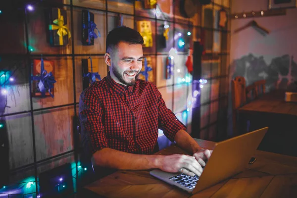 Thema opleiding en computers. Een jonge man met een baard en in een shirt maakt gebruik van een laptop, prints op het toetsenbord in een coffeeshop op een houten tafel in de avond. Kerst decor en hang een krans — Stockfoto