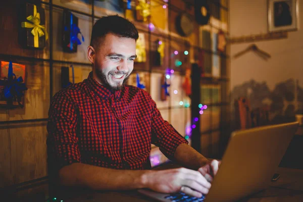 Entrenamiento temático y computadoras. Un joven con barba y camisa usa un portátil, impresiones en el teclado en una cafetería en una mesa de madera por la noche. Decoración de Navidad y colgar una guirnalda — Foto de Stock