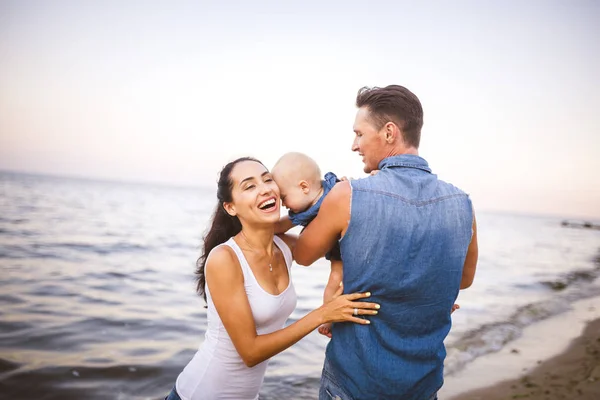 Hermosa familia joven de vacaciones con el bebé. El padre sostiene a la chica rubia en sus brazos, y la madre morena abraza a su marido. El concepto de unas vacaciones de playa en familia verano cerca del mar —  Fotos de Stock