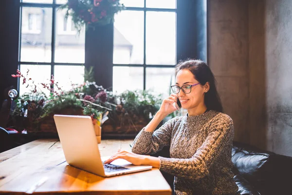Küçük işletme temasıdır. Bir dizüstü bilgisayar bir kafede Noel dekor ile dekore edilmiş ve telefonda konuşurken arkasında çalışan bir genç serbest kadın. Bir gri kazak ve gözlük giymiş — Stok fotoğraf