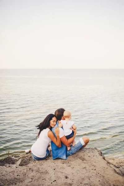 Tema vacaciones en familia con un niño pequeño en la naturaleza y el mar. Mamá, papá y la hija de un año están sentados en el abrazo, las niñas en las manos del hombre, con su espalda en la altura del acantilado con vistas al mar —  Fotos de Stock