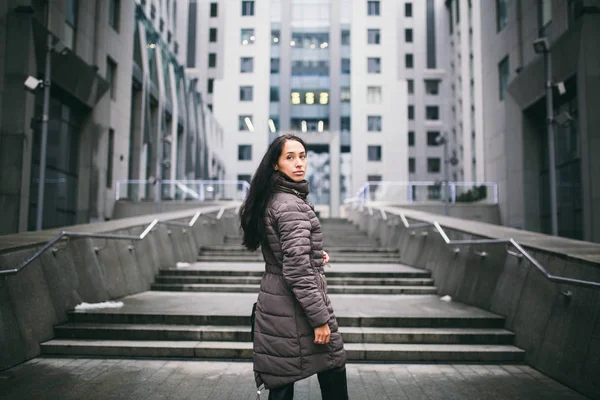 Retrato de uma mulher sobre um tema de negócios. Menina morena branca nova em jaqueta longa, casaco com saco de couro preto fica no fundo do centro de negócios, prédio de escritórios com fachada de vidro — Fotografia de Stock