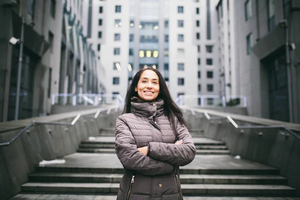 Retrato de una mujer sobre un tema de negocios. Joven morena caucásica con chica de pelo largo en chaqueta larga, abrigo se encuentra en el fondo del centro de negocios, edificio de oficinas con fachada de vidrio. Nublado en invierno —  Fotos de Stock