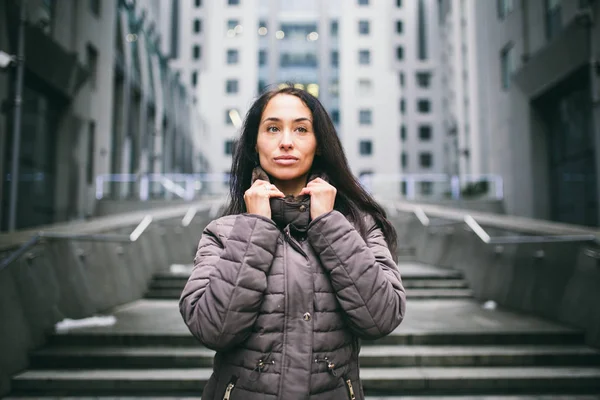 Portrait of a woman on a business theme. Young caucasian brunette with long hair girl in long jacket, coat stands on business center background, office building with glass facade. Overcast in winter — Stock Photo, Image